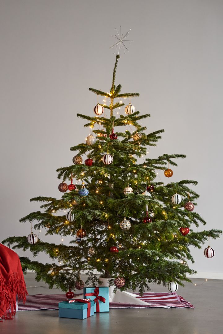 Dekorera julgranen med årets julgranspynt 2021 i 4 olika stilar enligt Nest Trends - Nurture, Share, Boost och Cultivate. Här ser du en färgstark och lekfull julgran på en randig matta från HAY i rött och vitt samt vit julgransfot från Born in Sweden.